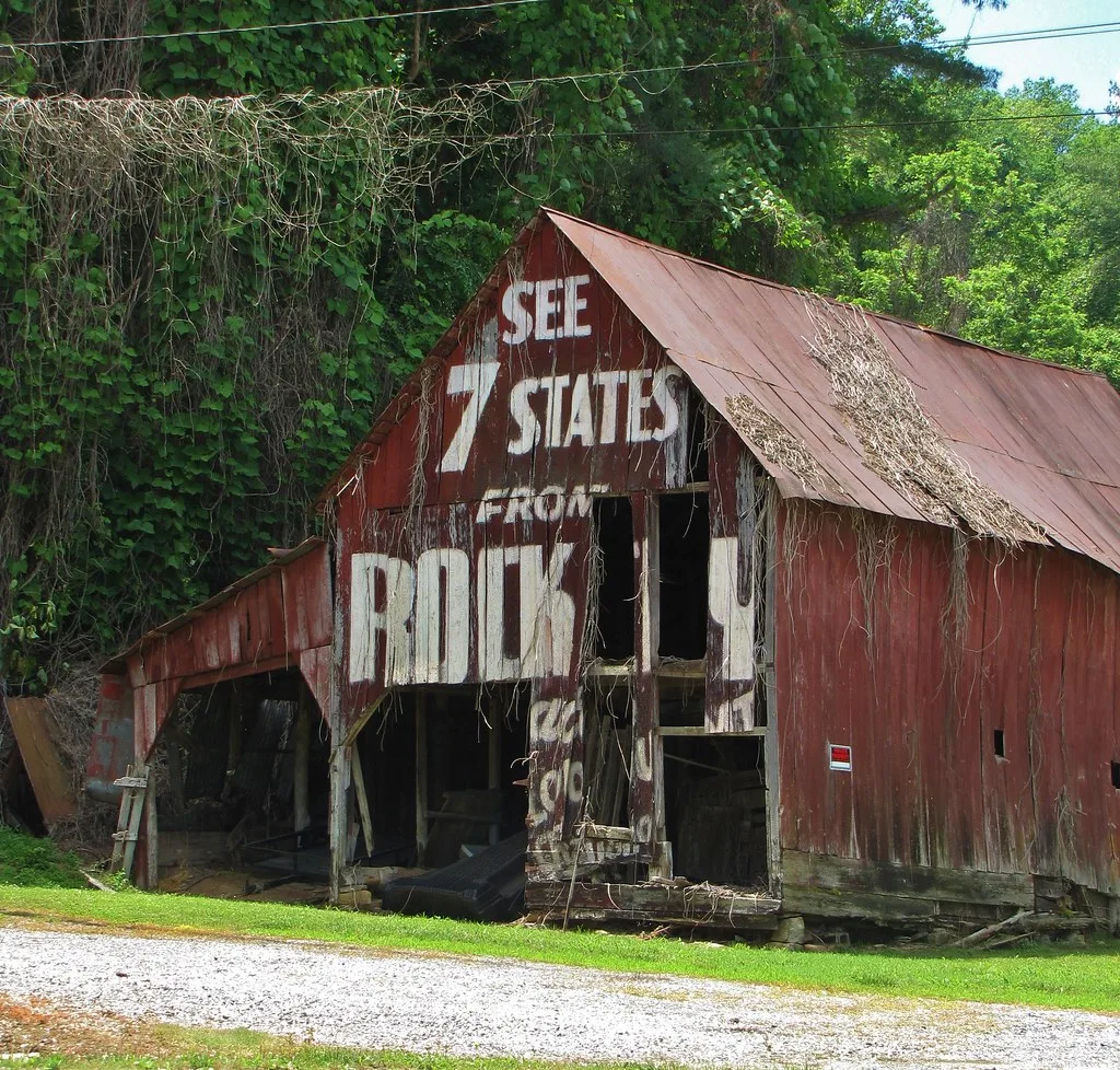 Ruined red barn bearing an old white advertisement, with large gaping openings.