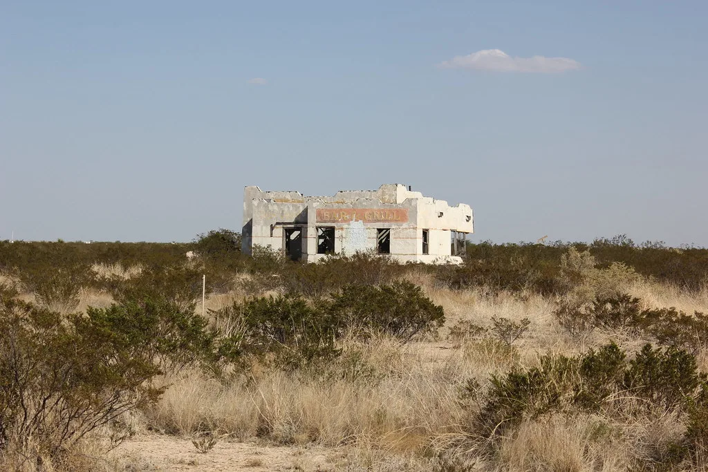 White ruin of an old bar standing among shrubs and dry grass.