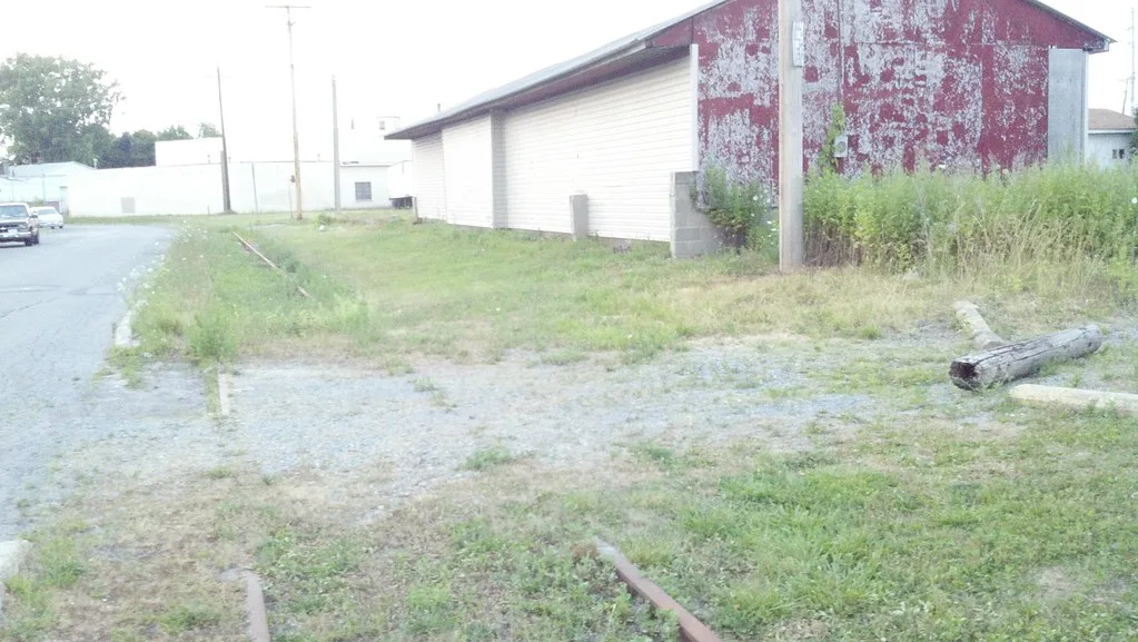 Remains of an abandoned railway track whose rusty rails disappear beneath gravel and grass beside a shed.