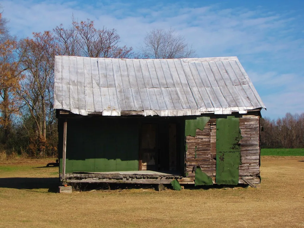 Small derelict wooden cabin with torn green cladding and a sagging porch beneath a metal roof.