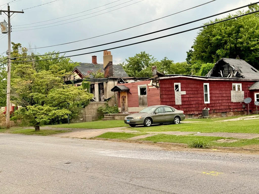 Maisons abandonnées à bardage rouge et murs noircis, aux toits effondrés et baies condamnées, derrière une voiture au bord de la rue.