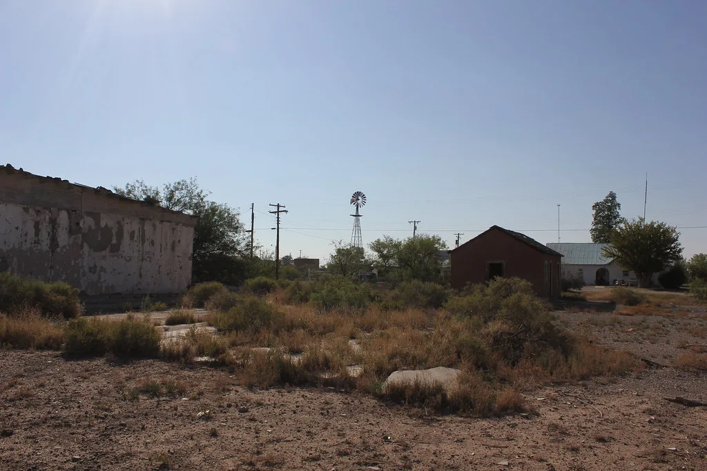 Abandoned buildings around a scrub-covered yard with a windmill in the background.