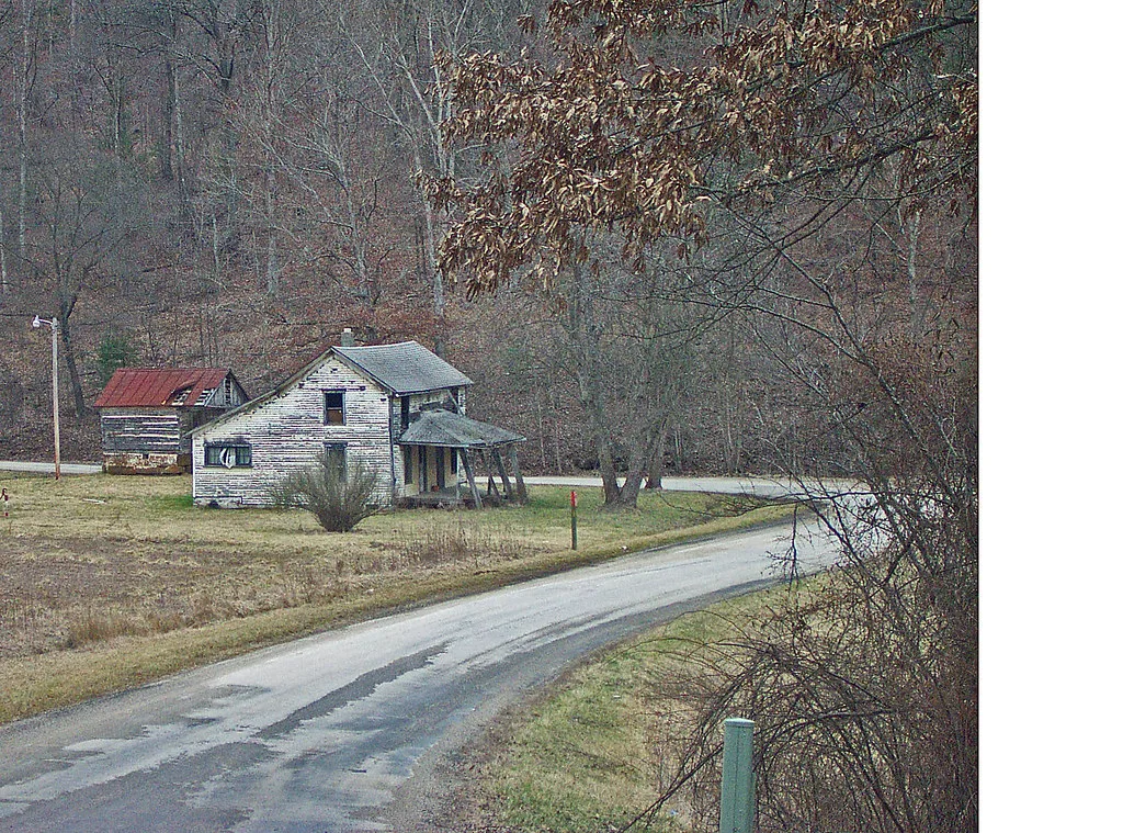 Maison de bois blanc vieillie près d’une route, avec porche incliné et grange au toit rouge troué.
