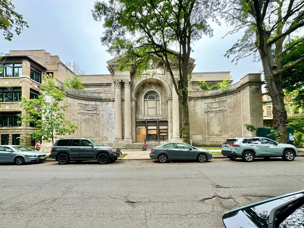Monumental stone facade with sealed doors and vegetation on its cornices behind parked cars.