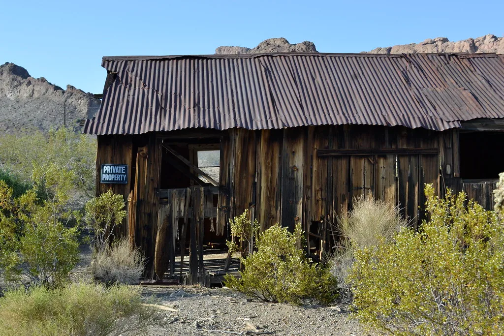 Dilapidated wooden cabin with a rusty metal roof in scrubland.
