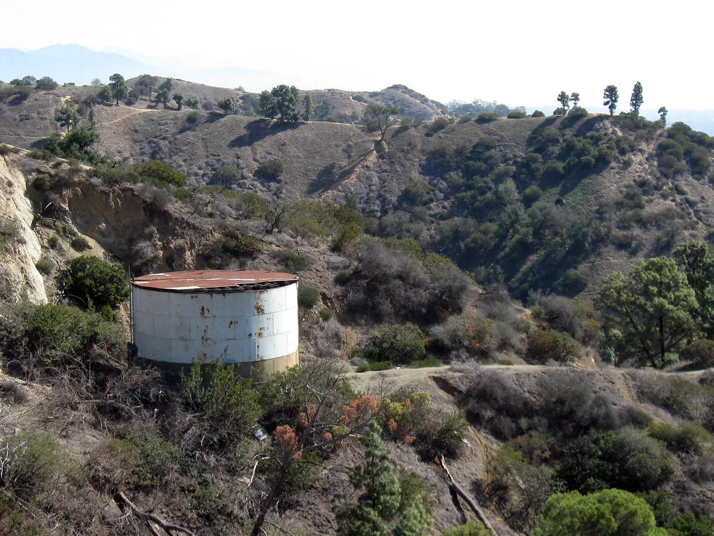 Ancien réservoir cylindrique rouillé à toit déchiré parmi les broussailles d’une colline sèche.