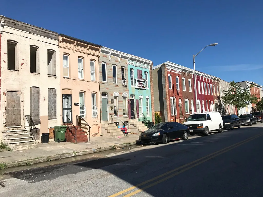 Maison abandonnée à façade claire et fenêtres vides au début d’une rangée de maisons colorées dans une rue.