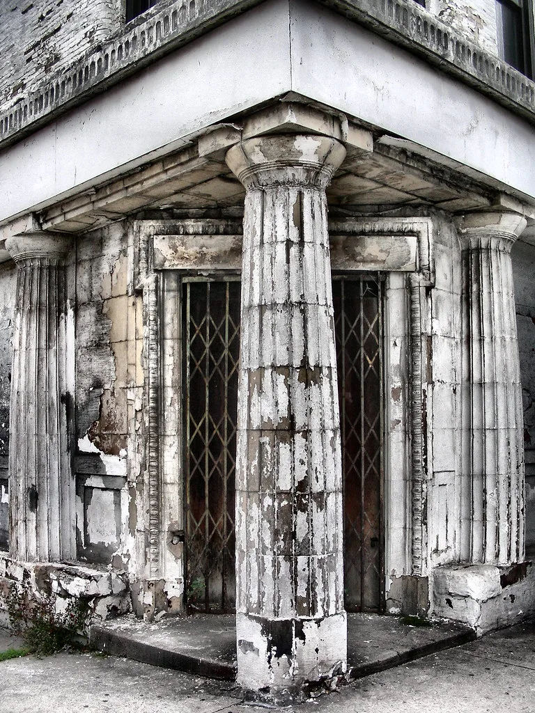 Corner porch with fluted columns and peeling paint in front of two gated doors.
