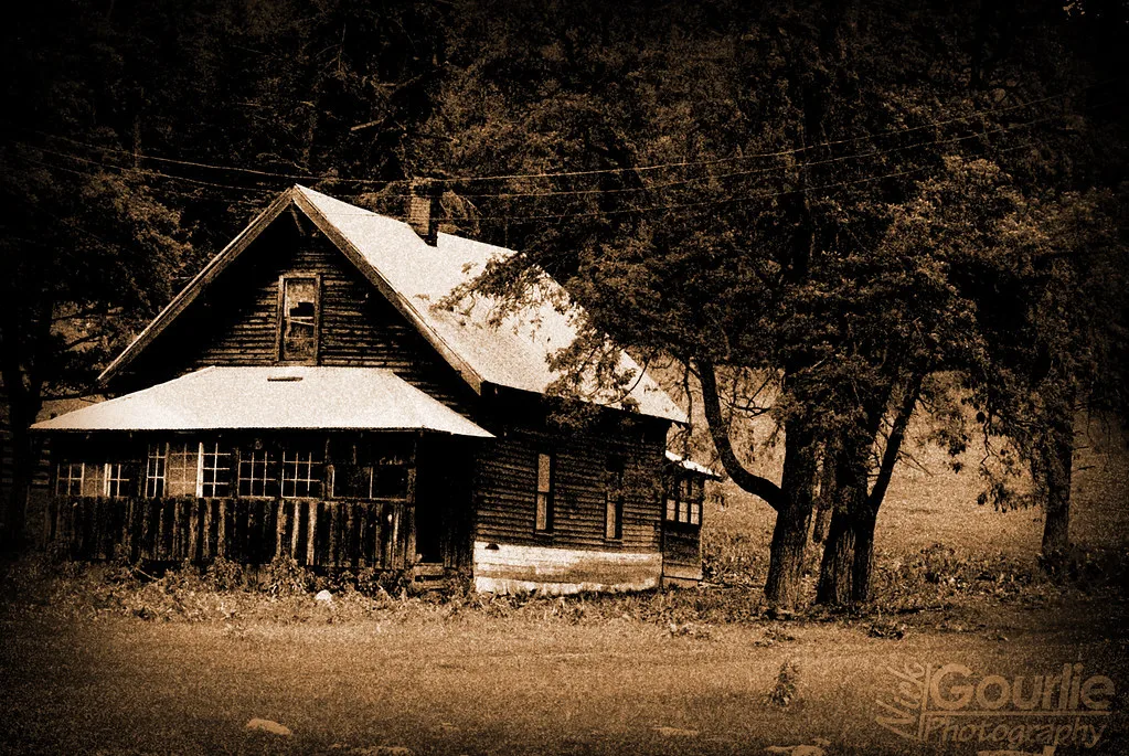 Abandoned dark wooden house with an enclosed porch with broken glazing beside large trees, in sepia.