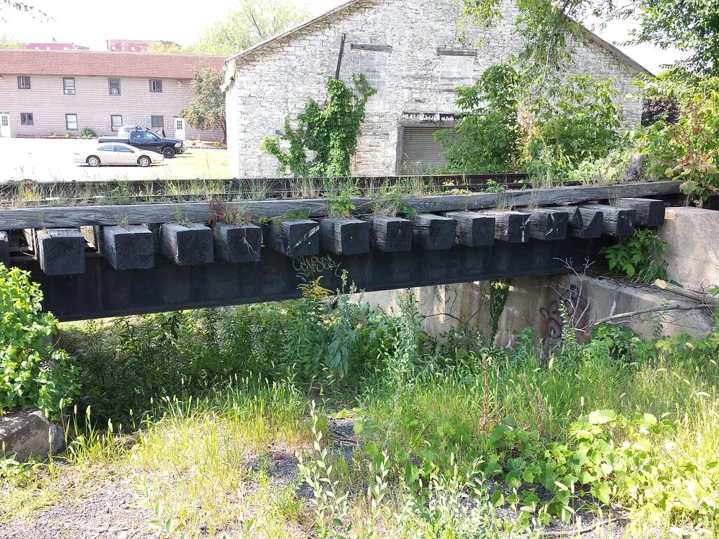 Ancien pont ferroviaire envahi d’herbes devant un bâtiment aux fenêtres murées.