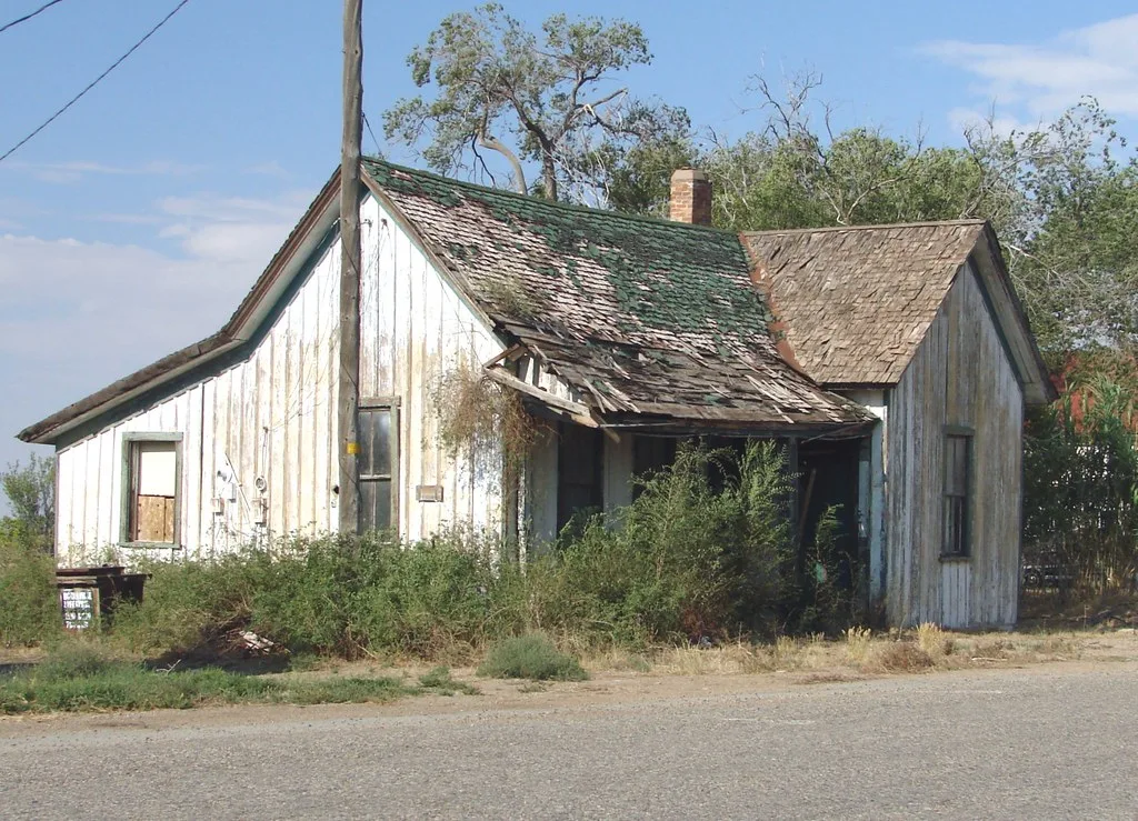 Small white wooden house with a damaged roof and a porch overgrown with scrub.