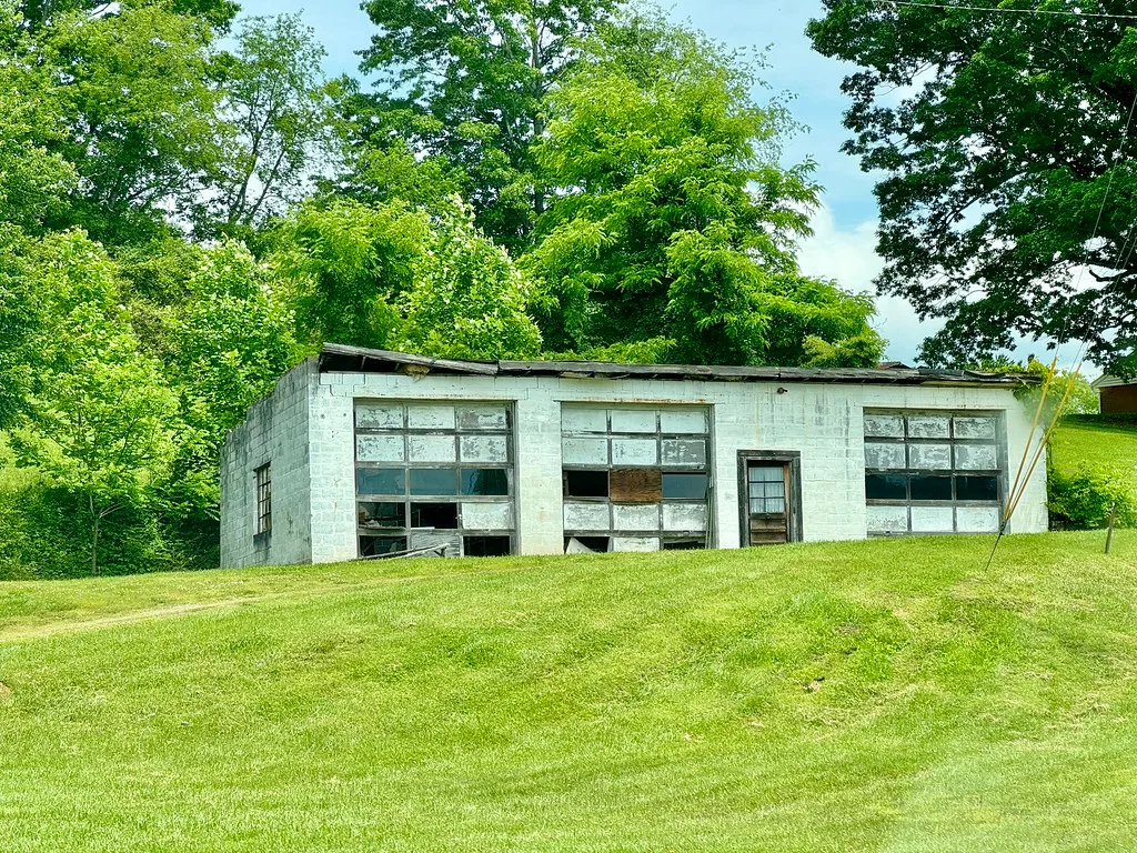 Old white garage with three deteriorated doors in front of trees.