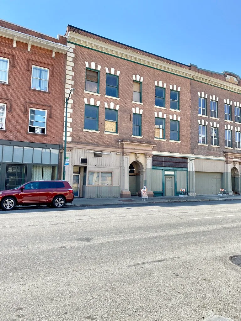 Ancien immeuble de briques aux fenêtres partiellement condamnées le long d’une rue.