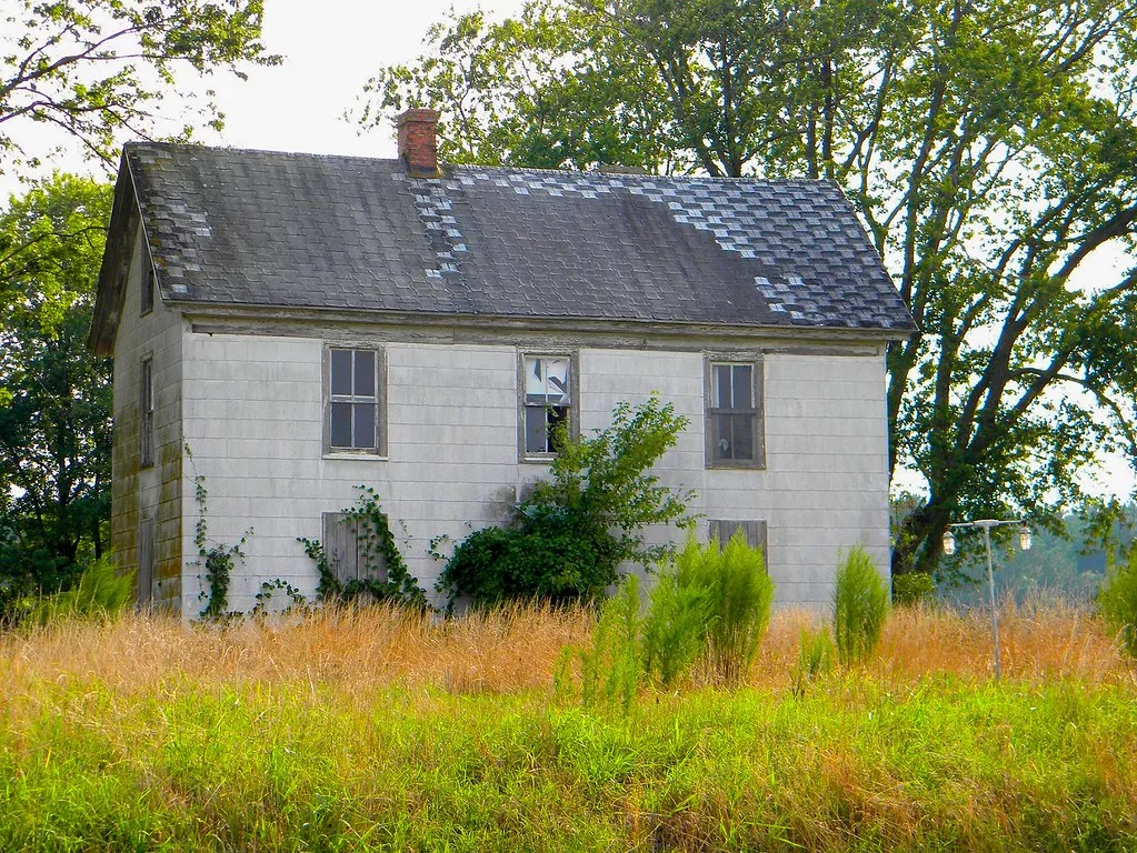 Maison blanche à deux niveaux, aux fenêtres cassées et condamnées, parmi les herbes dorées.