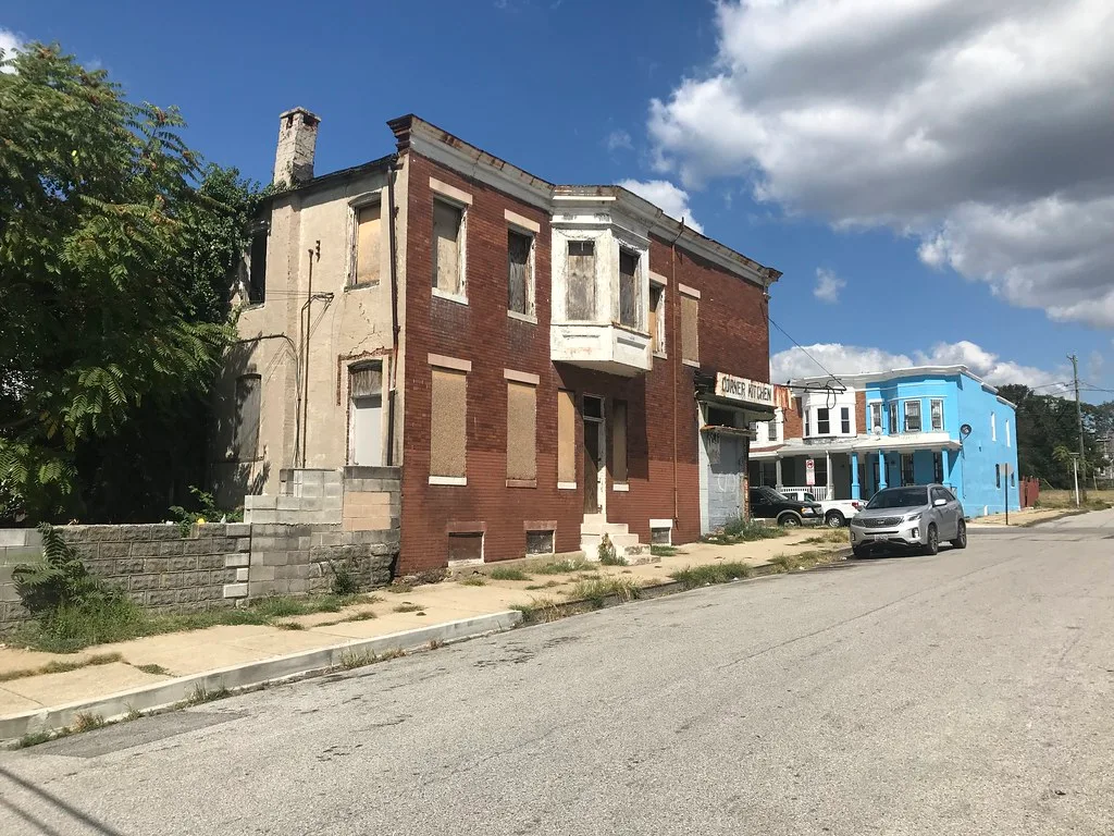 Maison abandonnée de brique rouge à baie en saillie blanche, toutes les fenêtres condamnées et enduit latéral fissuré.