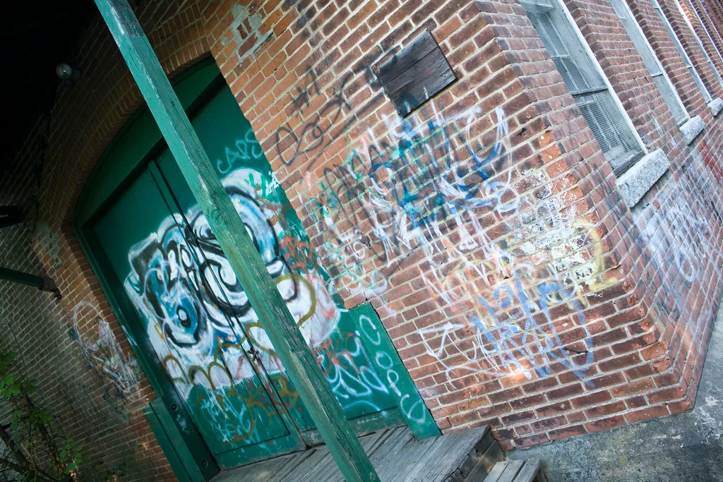 Graffiti-covered green door under the weathered wooden porch of a brick building.