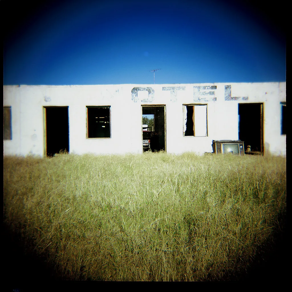 Abandoned white former motel with empty doorways and windows facing tall grass.