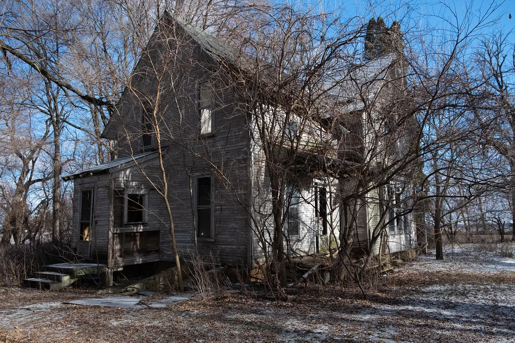 Large abandoned wooden house with broken windows and a sagging porch among bare trees.