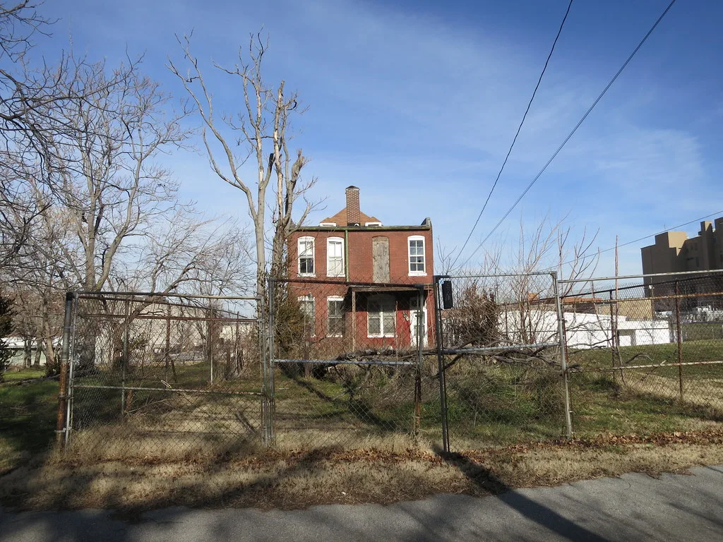 Two-storey red brick house with boarded openings behind wire fencing and fallen branches.