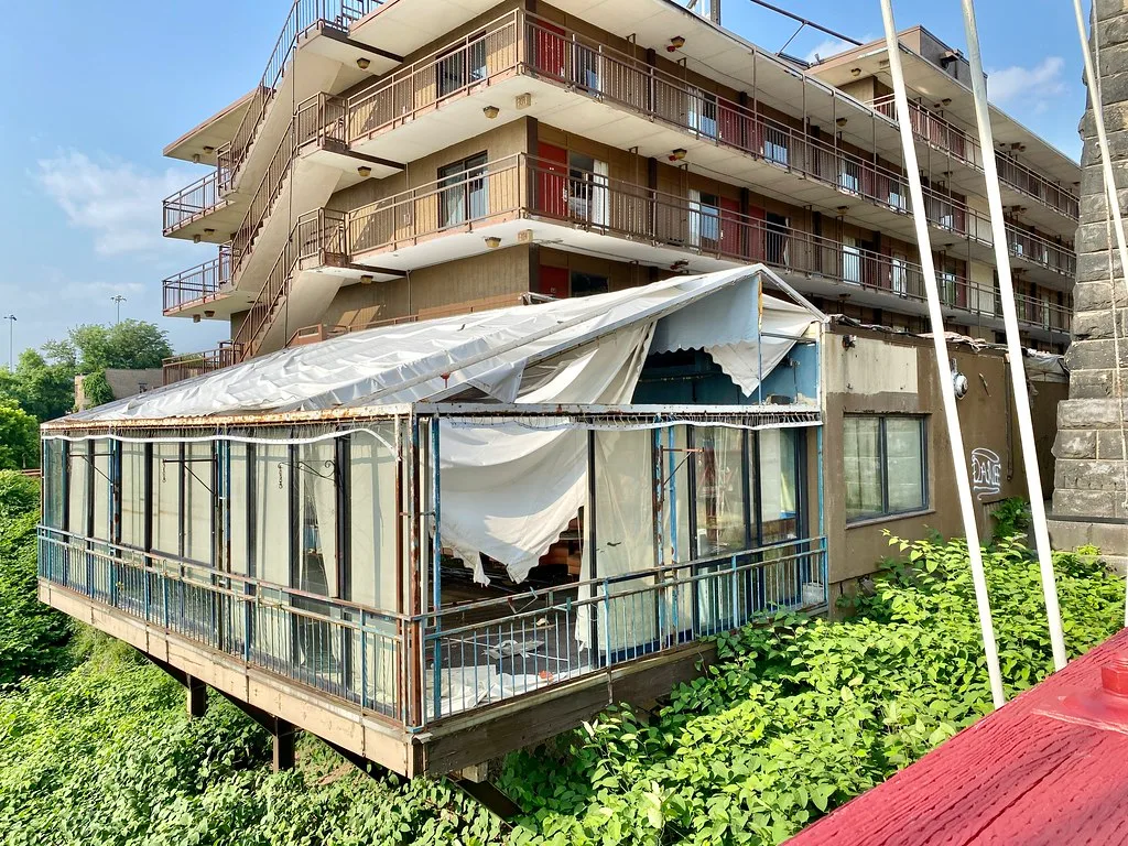 Abandoned veranda with missing glazing and torn roof fabric in front of a balconied building.