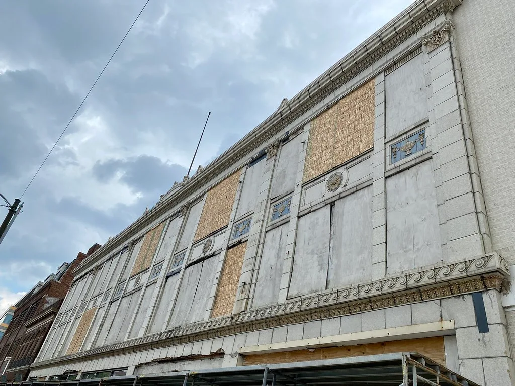 Old pale facade with large boarded openings beneath a cloudy sky.