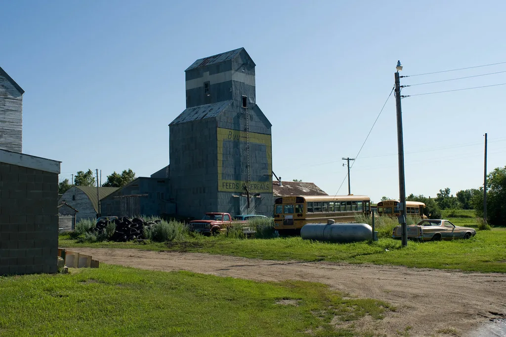 Ancien élévateur à grains abandonné aux tôles dégradées près de bus scolaires et voitures dans la végétation.