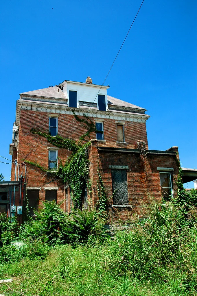 Old red-brick house with boarded windows in dense vegetation.