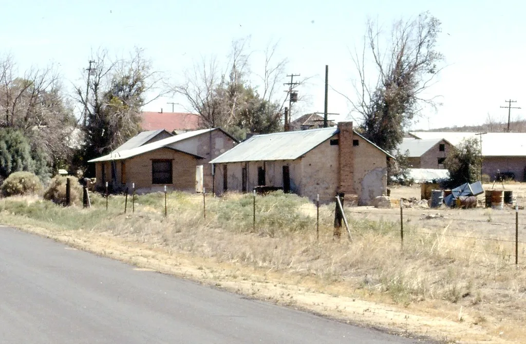 Petites maisons délaissées à toits métalliques et enduits écaillés derrière une clôture et des herbes sèches.