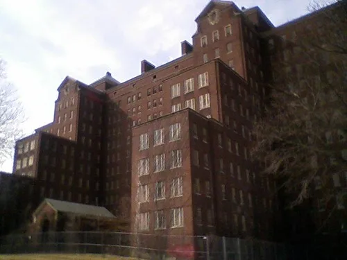 Large brick building with boarded windows behind a fence.