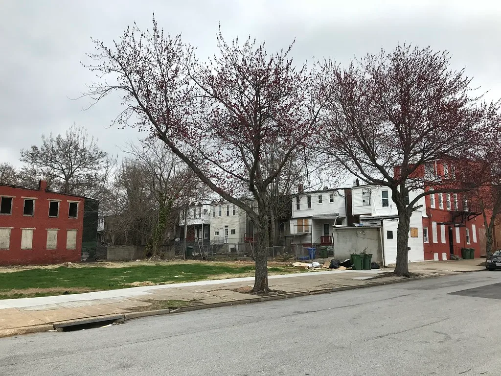 Small red abandoned building with empty upper windows and boarded lower openings beside an urban plot.
