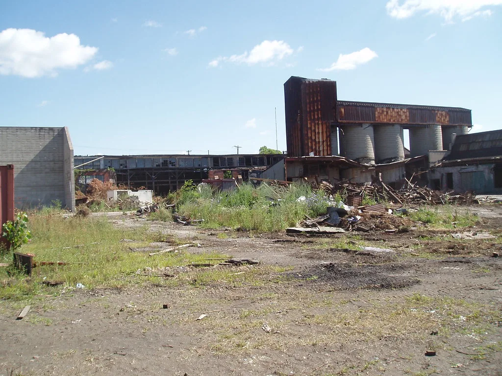 Old factory and rusty silos behind a yard overgrown with weeds and collapsed framing.
