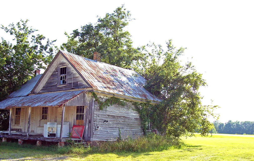 Small abandoned gray wooden house with a rusty metal roof, broken gable window and cluttered porch.
