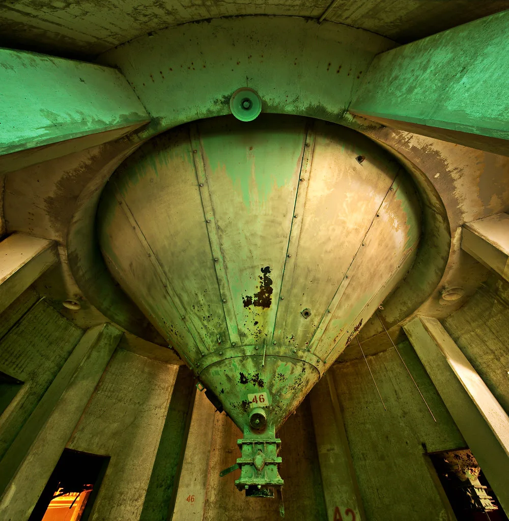 Large corroded green metal hopper between concrete pillars.