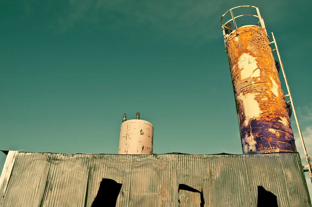 Old industrial building with torn metal siding beneath a heavily rusted cylindrical tower.