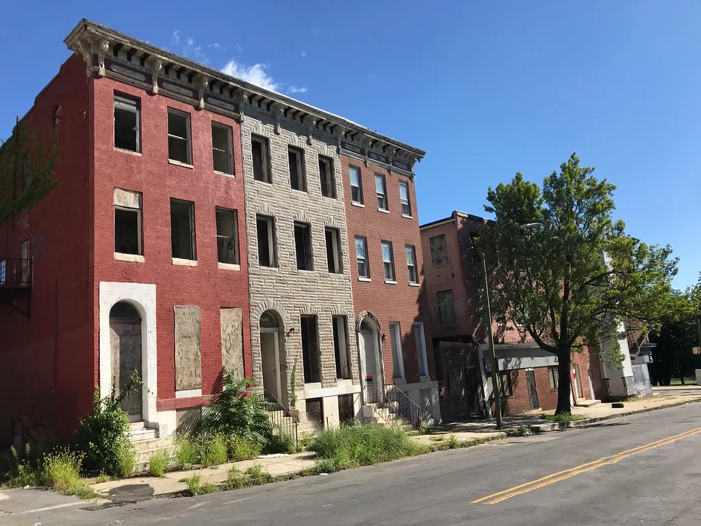 Abandoned three-storey red brick and pale stone houses with empty windows and boarded entrances lined with plants.