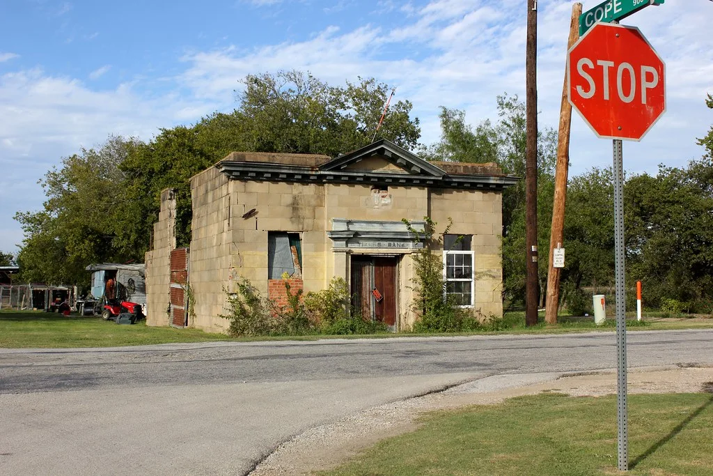 Facade of a small ruined bank with damaged openings and climbing plants beside an intersection.
