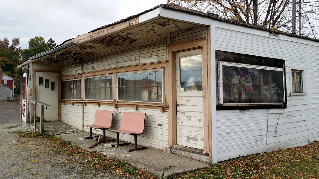 Small white shop with peeling paint and a decayed canopy, with two pink seats before the windows.