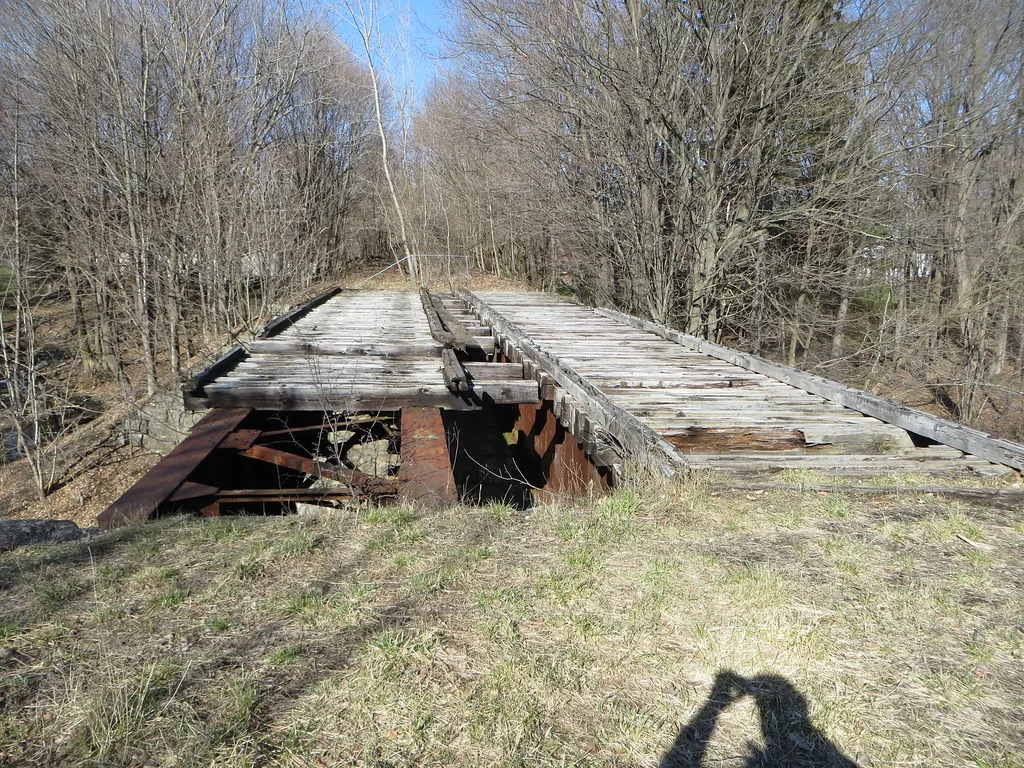Ancien pont à traverses de bois cassées laissant voir des poutres rouillées entre les arbres.