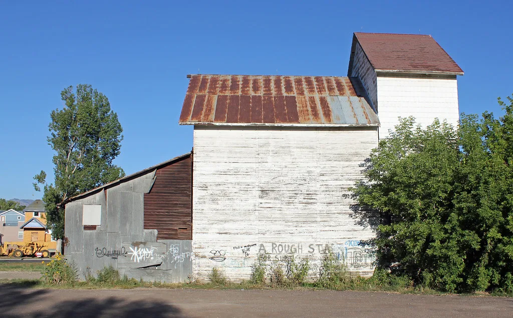 Old white building with a square tower, rusty roof and graffiti-covered walls overgrown with plants.