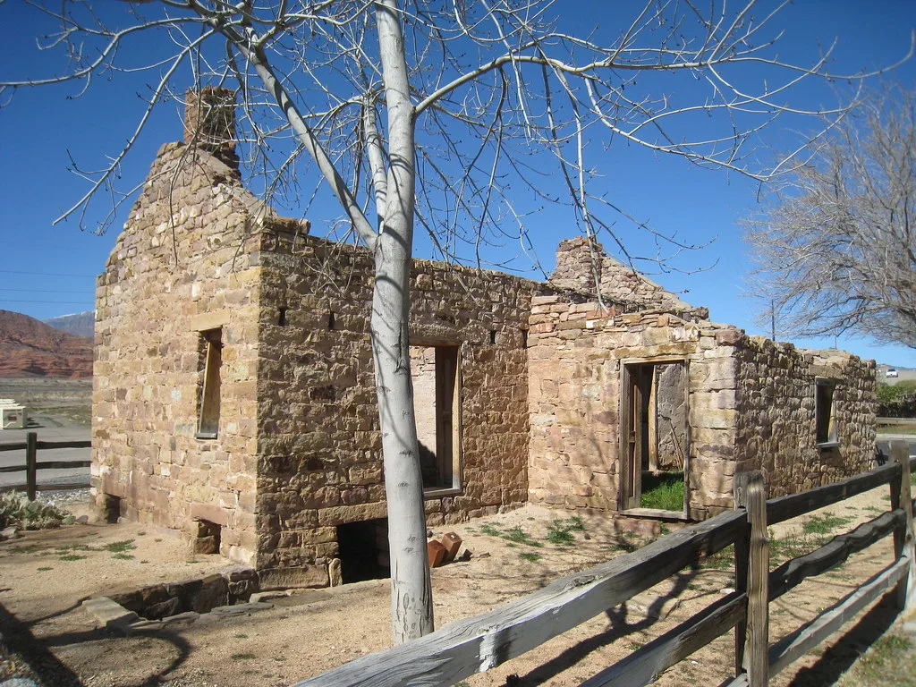 Ruine de maison en pierre avec cheminée et ouvertures vides derrière une barrière de bois.