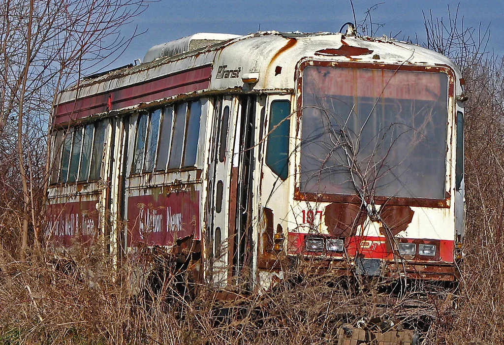 Old white and red tram with peeling paint surrounded by branches and dry grass.