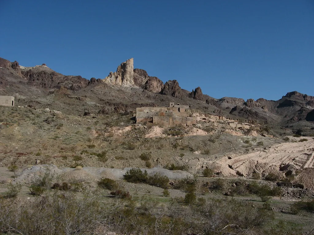 Concrete building ruins spread across a rocky desert slope beneath jagged peaks.