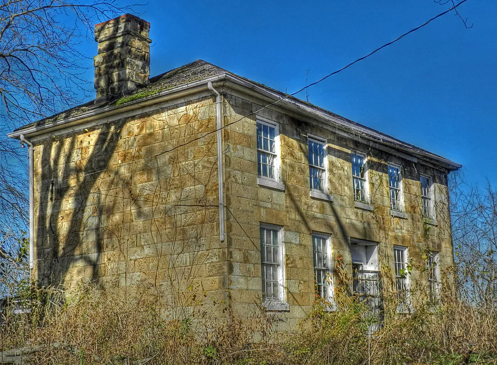 Abandoned yellow stone house with broken windows, surrounded by shrubs growing against its facade.