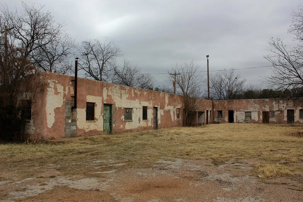 Old low complex with deteriorated render and empty openings around a grassy courtyard.