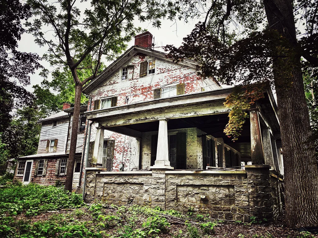 Grande maison abandonnée à porche de pierre, peinture écaillée et fenêtres brisées parmi les arbres.