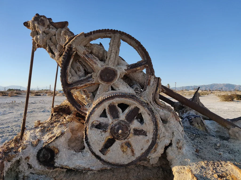 Large rusty gears of ruined machinery encrusted with pale deposits on arid ground.