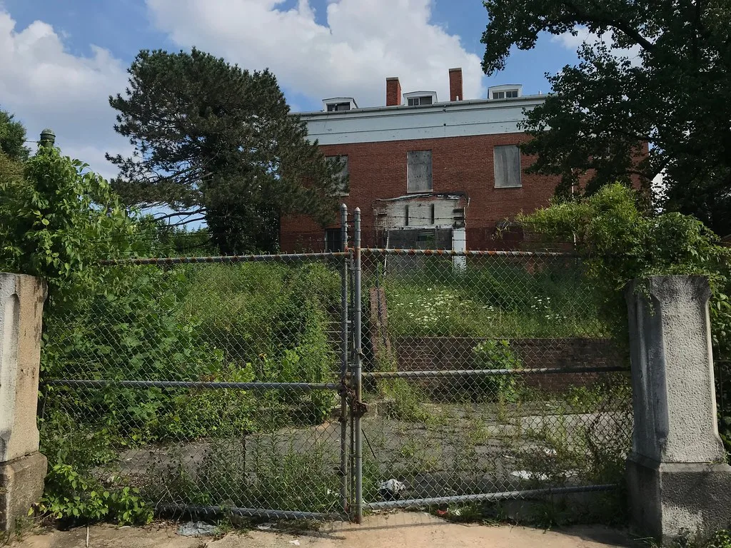 Brick building with boarded windows behind a rusty gate and tall weeds.