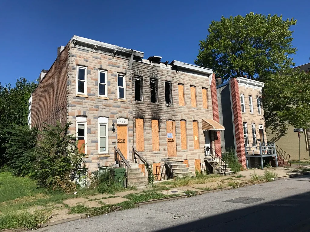 Rangée de maisons abandonnées à parement clair, dont une façade noircie par le feu avec fenêtres vides et portes condamnées.