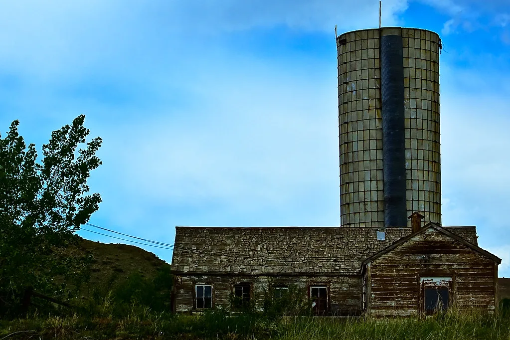 Old wooden farm building with broken windows in front of a tall cylindrical silo.