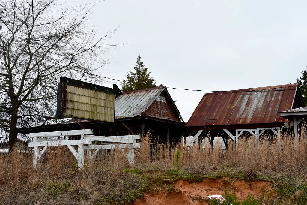 Old wooden buildings with rusty roofs and an empty sign in tall grass.